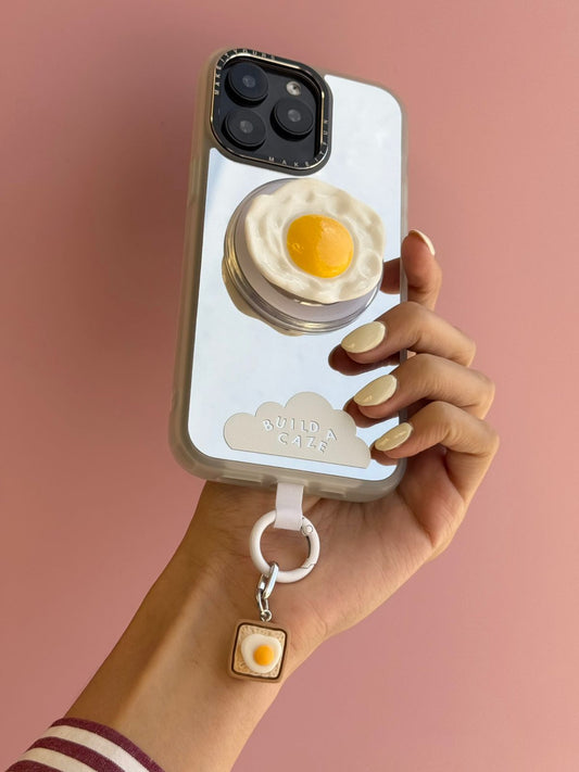 Un charm y accesorio colgante Eggie Toast Bites Charmie en color marrón y blanco sujeto a un celular - Build A Caze